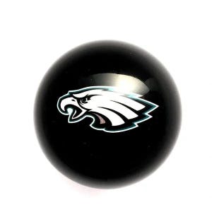 May include: A spherical, glossy black gear shift knob featuring a centered graphic of a white eagle head in profile, outlined in teal and black. The eagle design is oriented to the left, showcasing sharp, angular feathers and a distinct beak.