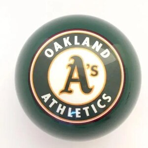 May include: A dark green spherical lawn bowl featuring a circular emblem in the center. The emblem has a white background with the text OAKLAND ATHLETICS in white capital letters and a large stylized gold and black A's logo in the middle.