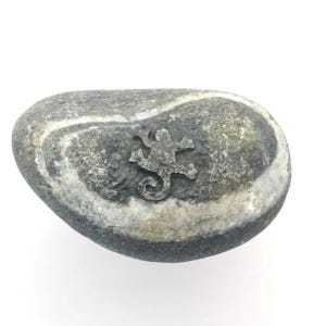 May include: A smooth, oval-shaped gray river stone featuring a natural white mineral band encircling the center. A small, dark, embossed lizard silhouette is centered on the surface of the stone, providing a textured contrast to the smooth rock.