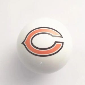 May include: A smooth, spherical white ball featuring the Chicago Bears logo printed in the center. The logo consists of a stylized orange letter C with a thick dark navy blue outline, centered on the surface of the round white object.