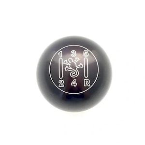 May include: A spherical black gear shift knob featuring a white engraved manual transmission pattern. The pattern displays the numbers 1, 2, 3, 4, 5, and R, with a central white line drawing of a gecko silhouette positioned between the gear numbers.