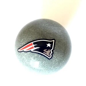 May include: A spherical object with a silver glitter finish featuring a dark blue, red, and white logo of a stylized profile of a person wearing a tricorne hat with a white star, representing the New England Patriots football team emblem.