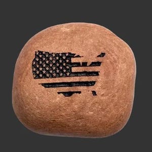 May include: A smooth, rounded tan river stone featuring a black laser-engraved design of the United States map filled with the pattern of the American flag, including stars in the upper left corner and horizontal stripes across the landmass.