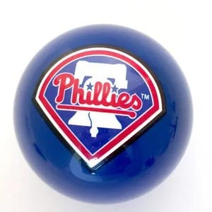 May include: A smooth, spherical blue object featuring a centered graphic of the Philadelphia Phillies logo. The logo consists of a white Liberty Bell silhouette inside a diamond shape with red and white borders and the red text Phillies.