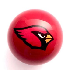 May include: A smooth, spherical red object featuring a graphic of a cardinal bird head in profile. The bird design is rendered in black, white, and yellow, centered on the glossy red surface of the round item, creating a bold, high-contrast visual.