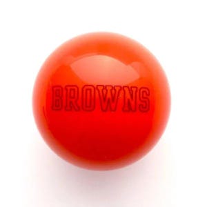 May include: A smooth, spherical, bright orange object with a glossy finish. The word BROWNS is embossed in capital letters across the center of the sphere in a matching orange tone, creating a subtle, monochromatic, and textured appearance.