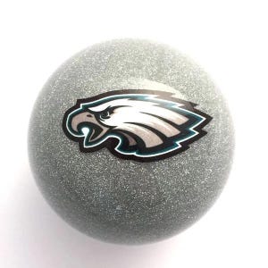 May include: A spherical object with a silver glitter finish featuring a graphic of a white eagle head with black and teal outlines. The eagle is depicted in profile facing left with an open beak, centered on the round, textured surface.