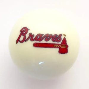 May include: A smooth, off-white baseball featuring the red and blue Atlanta Braves logo printed on the front. The logo includes the word Braves in a stylized script font with a red tomahawk graphic underneath, complete with a small trademark symbol.