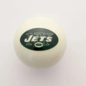 May include: A smooth, spherical white ball featuring a dark green oval logo in the center. The logo displays the letters NY in a bold outline font, the word JETS in white capital letters, and a small green football icon positioned below the text.