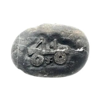 May include: A smooth, dark gray oval stone featuring a carved, recessed design of a four-wheeled off-road vehicle. The carving shows the silhouette of a jeep-style truck with two prominent circular wheels and a small, stylized emblem between them.