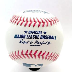 May include: A white baseball with red stitching featuring the Rawlings logo at the top, the text OFFICIAL MAJOR LEAGUE BASEBALL with blue stars, the signature of Robert D. Manfred Jr. as Commissioner, and the blue MLB logo at the bottom.