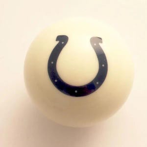 May include: A smooth, off-white spherical object featuring a dark blue horseshoe graphic centered on its surface. The horseshoe design includes small white dots along its perimeter, creating a distinct contrast against the light-colored ball.