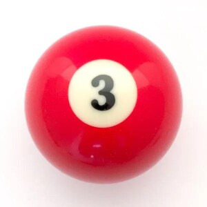 May include: A smooth, spherical billiard ball with a glossy red finish. Centered on the ball is a white circular inlay featuring the number 3 printed in a bold, black sans-serif font. The surface reflects light, indicating a hard, polished material.