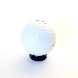 May include: A white golf ball with a dimpled surface texture mounted on a black cylindrical base. The golf ball features a small red number printed on its side. The base is solid black and includes a visible circular opening on its side.