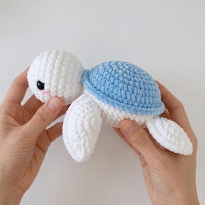 Può includere: Una piccola tartaruga di peluche lavorata all'uncinetto a mano, con testa e corpo bianchi e un guscio testurizzato azzurro. La tartaruga presenta due occhi di sicurezza in plastica nera e piccoli dettagli rosa ricamati sulle guance.