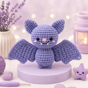 Puede incluir: Un murciélago de ganchillo morado con grandes orejas puntiagudas, ojos negros, una pequeña nariz rosa y dos colmillos blancos. El peluche tiene las alas extendidas y un cuerpo redondo, elaborado con un punto de hilo texturizado en color lavanda.