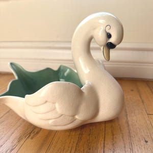 May include: A ceramic swan-shaped dish featuring a cream-colored exterior with a fine crackle glaze finish. The swan has a curved neck, a black eye detail, and a light yellow beak. The interior of the dish is a smooth, solid sage green color.