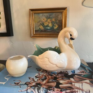 May include: A cream-colored ceramic swan-shaped bowl with a light green interior, accompanied by a small, round, cream-colored ceramic vessel. Both items are positioned on a book titled Los Angeles with a photographic cover featuring people.