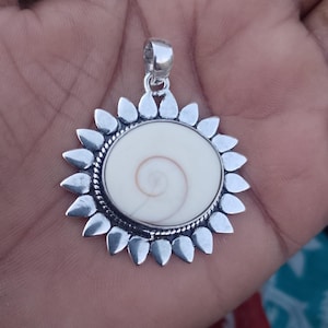 May include: A circular pendant featuring a central cream-colored Shiva eye shell with a natural spiral pattern, encased in a silver-toned metal setting with a rope-textured border and a sunburst design of pointed petals, topped with a silver bail.