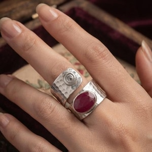 May include: A wide silver ring featuring a hammered texture, a spiral design element, and an oval-shaped deep red gemstone set into the metal band. The ring has an open-ended design that wraps around the finger with a polished finish.