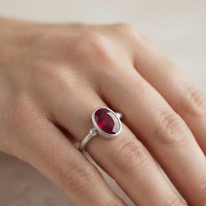 May include: A silver ring featuring a central oval-cut deep red gemstone set in a smooth bezel. The band has a matte finish and includes a small spherical accent on each side of the stone setting, creating a clean and balanced design.