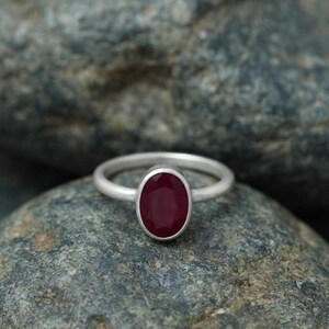 May include: A silver ring featuring a single oval-shaped, deep red gemstone set in a smooth metal bezel. The band of the ring has a matte, brushed finish, and the gemstone is faceted, reflecting light across its dark crimson surface.