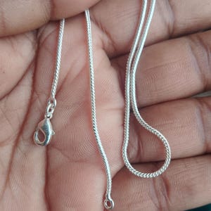 May include: A silver-coloured snake chain necklace featuring a polished finish and a lobster claw clasp. The chain has a smooth, rounded, and flexible texture with a continuous woven pattern throughout its length, ending in a small circular jump ring.