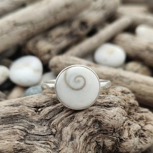 Puede incluir: Un anillo de plata con una incrustación de piedra circular de color blanco hueso que presenta un patrón de espiral marrón natural en el centro. La banda es lisa y metálica, sujetando el engaste redondo con un sencillo borde de plata pulida.