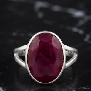 May include: A silver ring featuring a large, oval-shaped, faceted deep red gemstone set in a smooth bezel. The band of the ring is split into two thin, polished silver segments that meet at the sides to support the central stone setting.