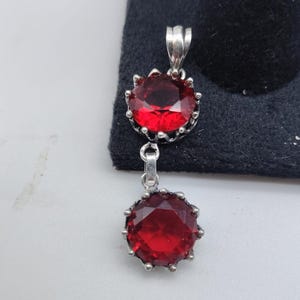 Puede incluir: Un colgante de plata con dos piedras rojas redondas y facetadas en engastes de garras. Las piedras están dispuestas verticalmente, con la superior conectada a una anilla y la inferior suspendida de la superior mediante un eslabón.