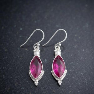 Puede incluir: Un par de pendientes colgantes de plata con piedras preciosas de color rosa vibrante en talla marquesa. Cada pendiente tiene un borde de plata texturizado tipo cuerda, un pequeño detalle de cuenta de plata y un gancho curvo.