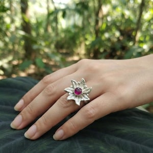 Può includere: Un anello in argento con un design a forma di fiore con petali testurizzati e una singola pietra preziosa rotonda di colore rosa intenso incastonata al centro. Il metallo ha una finitura lucida e i petali si irradiano dalla pietra.