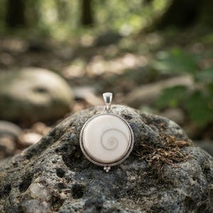 Puede incluir: Un colgante circular que presenta una concha de Shiva de color crema suave con un patrón de espiral marrón natural. La concha está encerrada en un marco de metal plateado pulido con un borde de cuerda trenzada y una pequeña cuenta decorativa.