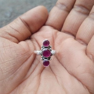 May include: A silver ring featuring three round, faceted, deep pink stones arranged vertically. The central stone is the largest, flanked by two smaller stones. The metal band displays intricate decorative metalwork with small bead accents.