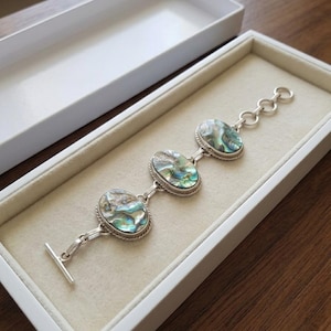 May include: A silver-toned link bracelet featuring three oval-shaped abalone shell cabochons with iridescent blue and green hues. The bracelet includes a toggle clasp closure and a short chain extension with circular links, presented in a white box.