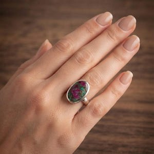 Puede incluir: Un anillo de plata con una piedra de forma ovalada y textura moteada en tonos magenta intenso, verde oscuro y negro. La piedra está engastada en una banda de plata pulida y lisa que rodea el dedo, mostrando una estética mineral natural.