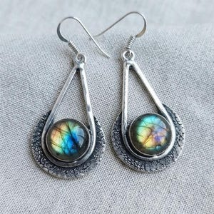May include: A pair of silver-toned drop earrings featuring a teardrop-shaped frame and a circular base with a textured finish. Each earring contains a round, iridescent labradorite gemstone displaying flashes of blue, yellow, and green colours.