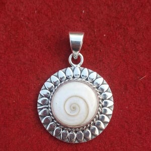 May include: A circular pendant featuring a central white Shiva eye shell with a natural spiral pattern. The shell is encased in a twisted metal border, surrounded by a decorative frame of polished silver-toned petal shapes and a top bail.