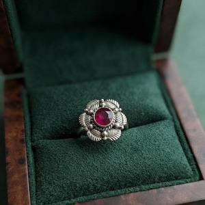 May include: A silver ring featuring a central round faceted deep red gemstone. The setting is designed with a series of textured silver leaves arranged in a floral pattern around the stone, accented by small silver beads between the petals.