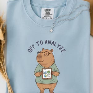 Puede incluir: Una camiseta azul claro de cuello redondo con un gráfico de un capibara marrón que lleva gafas redondas y una camisa verde. El capibara sostiene una tableta con gráficos coloridos. El texto sobre el gráfico dice OFF TO ANALYZE en azul oscuro.