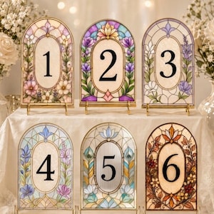 Puede incluir: Un conjunto de seis números de mesa de vitral en forma de arco, numerados del 1 al 6, con motivos florales y botánicos en varios colores. Cada pieza se sostiene sobre un soporte de metal dorado. El texto dice: STAINED GLASS TABLE NUMBERS + BUNDLE OF 6 DESIGNS +.