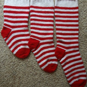 May include: Three red and white striped Christmas stockings. The stockings are knit with a red cuff and toe, and white and red horizontal stripes. The stockings are arranged on a neutral-colored carpet.