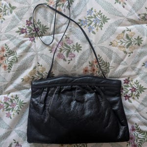 May include: A black leather handbag featuring a soft, gathered texture on the front panel, a smooth top trim with a subtle wave-like edge, and a thin, long shoulder strap attached to the sides for carrying over the shoulder or across the body.