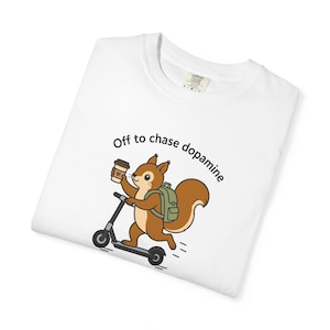May include: A white t-shirt featuring a cartoon illustration of a brown squirrel wearing a green backpack, riding a black scooter, and holding a coffee cup. The text above the squirrel reads, Off to chase dopamine, in a simple black font.