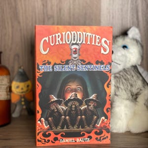 May include: A paperback book titled Curioddities: The Silent Sentinels by Daniel Bautz. The orange cover features an illustration of three figures with hats covering their eyes, ears, and mouths, positioned below a central eye graphic.