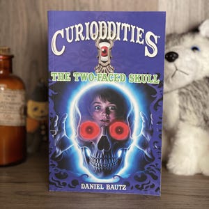 May include: A paperback book titled Curioddities: The Two-Faced Skull by Daniel Bautz. The cover features a blue background with a central illustration of a human skull with glowing red eyes, surrounding a child's face, and a keyhole graphic.