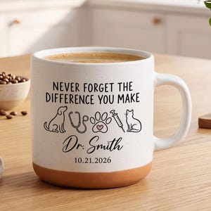 Puede incluir: Una taza de cerámica blanca con base de terracota que presenta el texto NEVER FORGET THE DIFFERENCE YOU MAKE, Dr. Smith, y 21.10.2026. El diseño incluye dibujos lineales negros de un perro, estetoscopio, huella con corazón rosa, jeringa y gato.