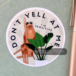 May include: A round sticker with the text "DON'T YELL AT ME I'M SENSITIVE" and an illustration of a hand with sunglasses and a potted plant. The sticker has a white background with a light green border.