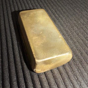 May include: A rectangular, solid brass ingot with a smooth, matte, metallic gold-toned surface. The object features rounded edges and a slightly weathered, uniform finish across its top and sides, presenting a heavy, block-like appearance.