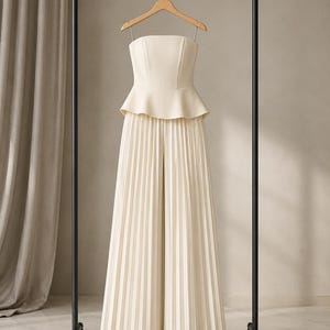 May include: A cream-colored strapless jumpsuit featuring a fitted bodice with a peplum waist and wide-leg trousers with vertical pleats throughout. The garment is displayed on a wooden hanger suspended from a black metal clothing rack.